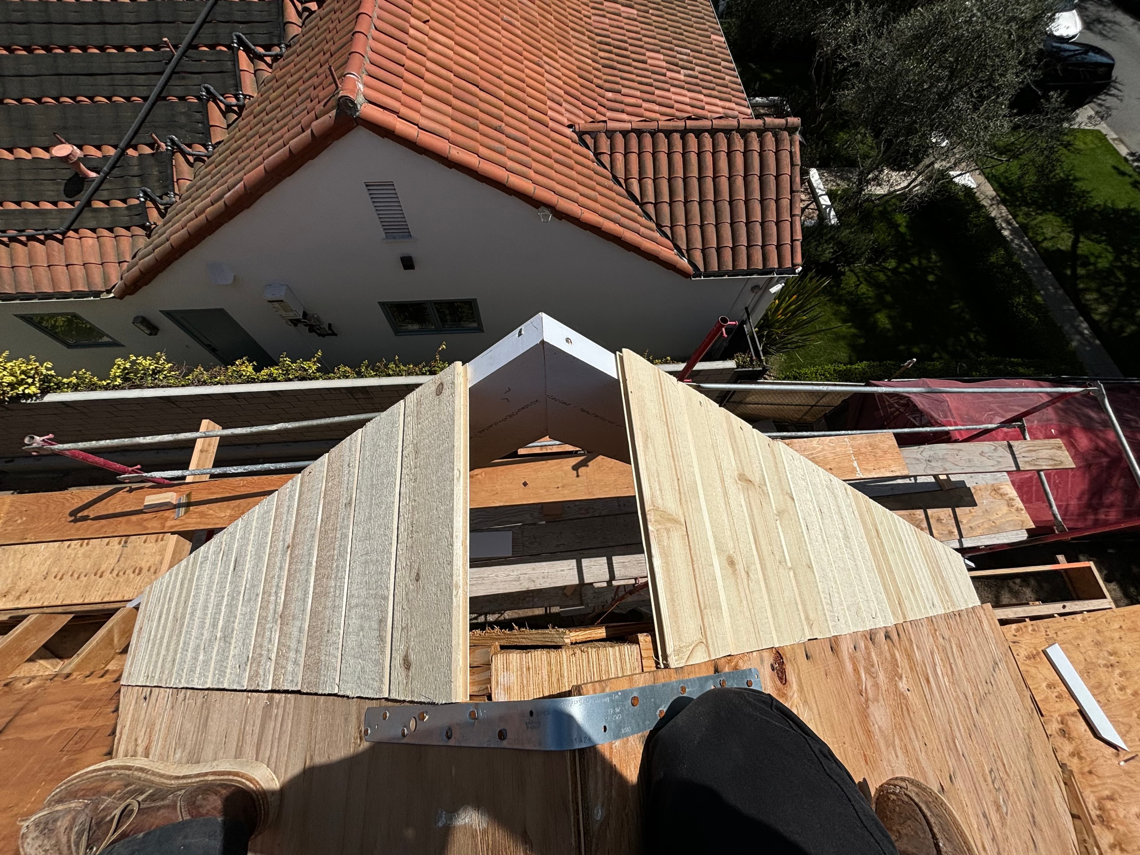 Wooden roof gable under construction on scaffolding, overlooking a house with a red tile roof.