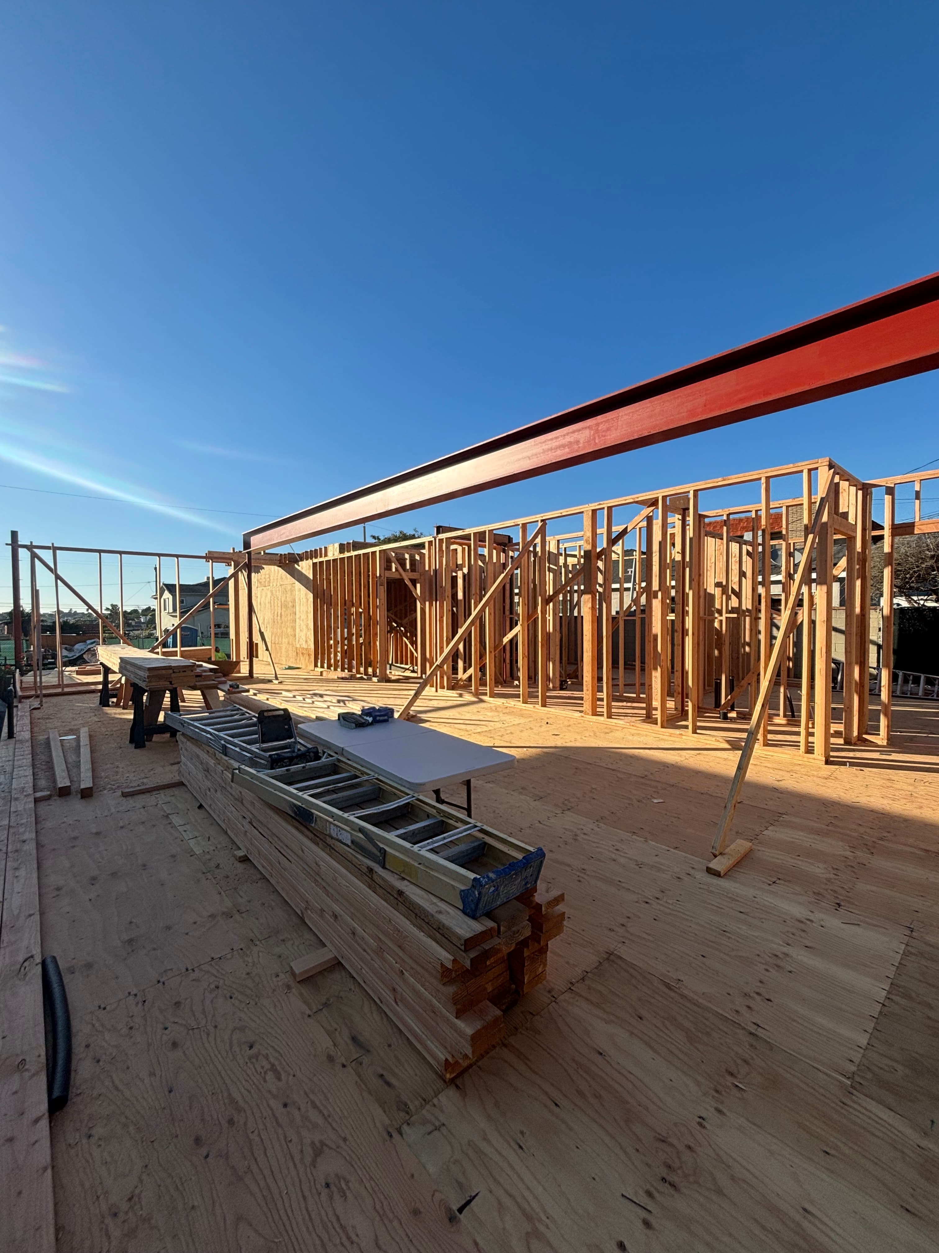Wooden house frame under construction with a prominent red steel beam under blue sky.