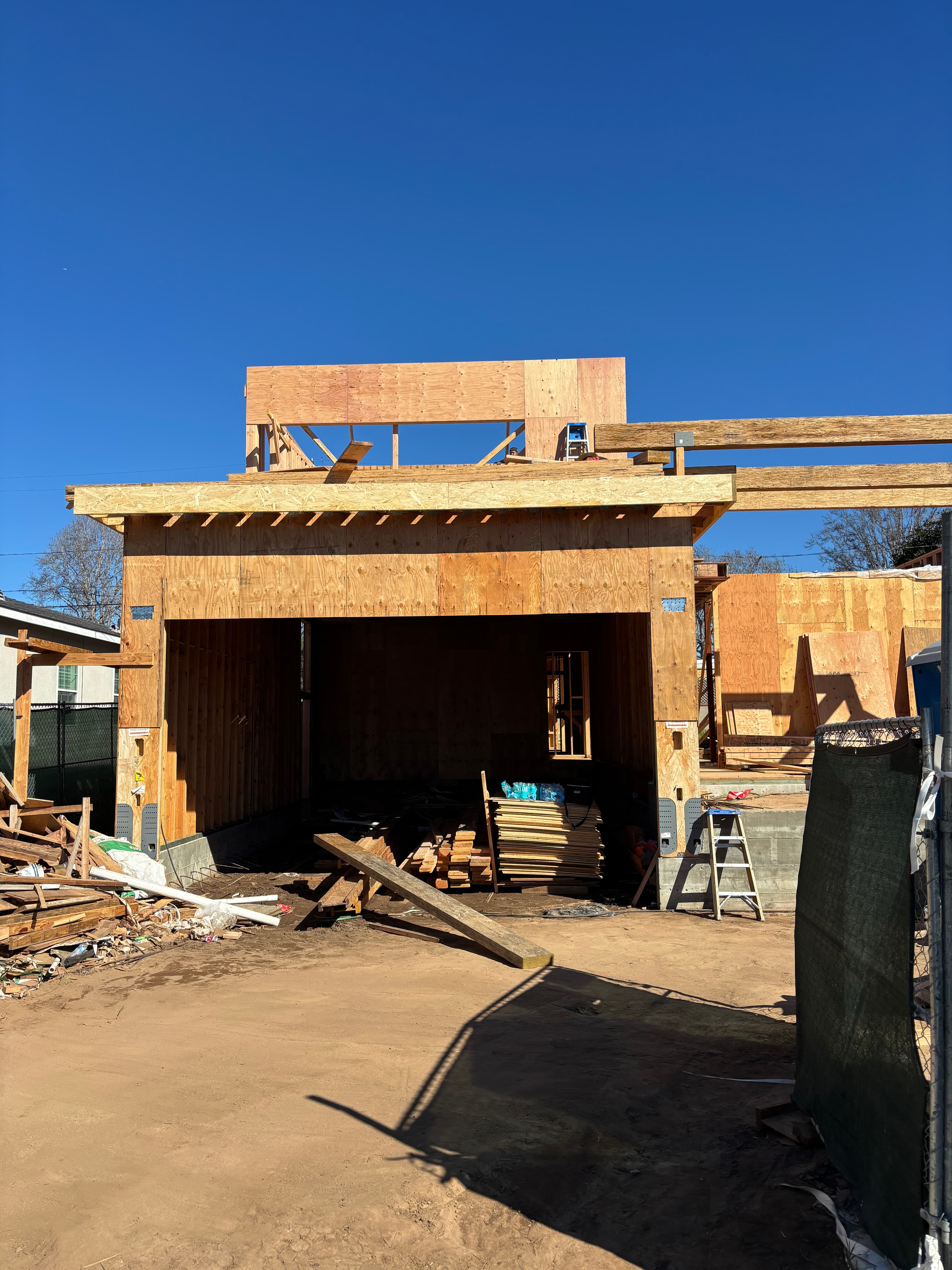 Wooden house frame under construction with plywood sheathing and lumber on a sunny dirt lot.