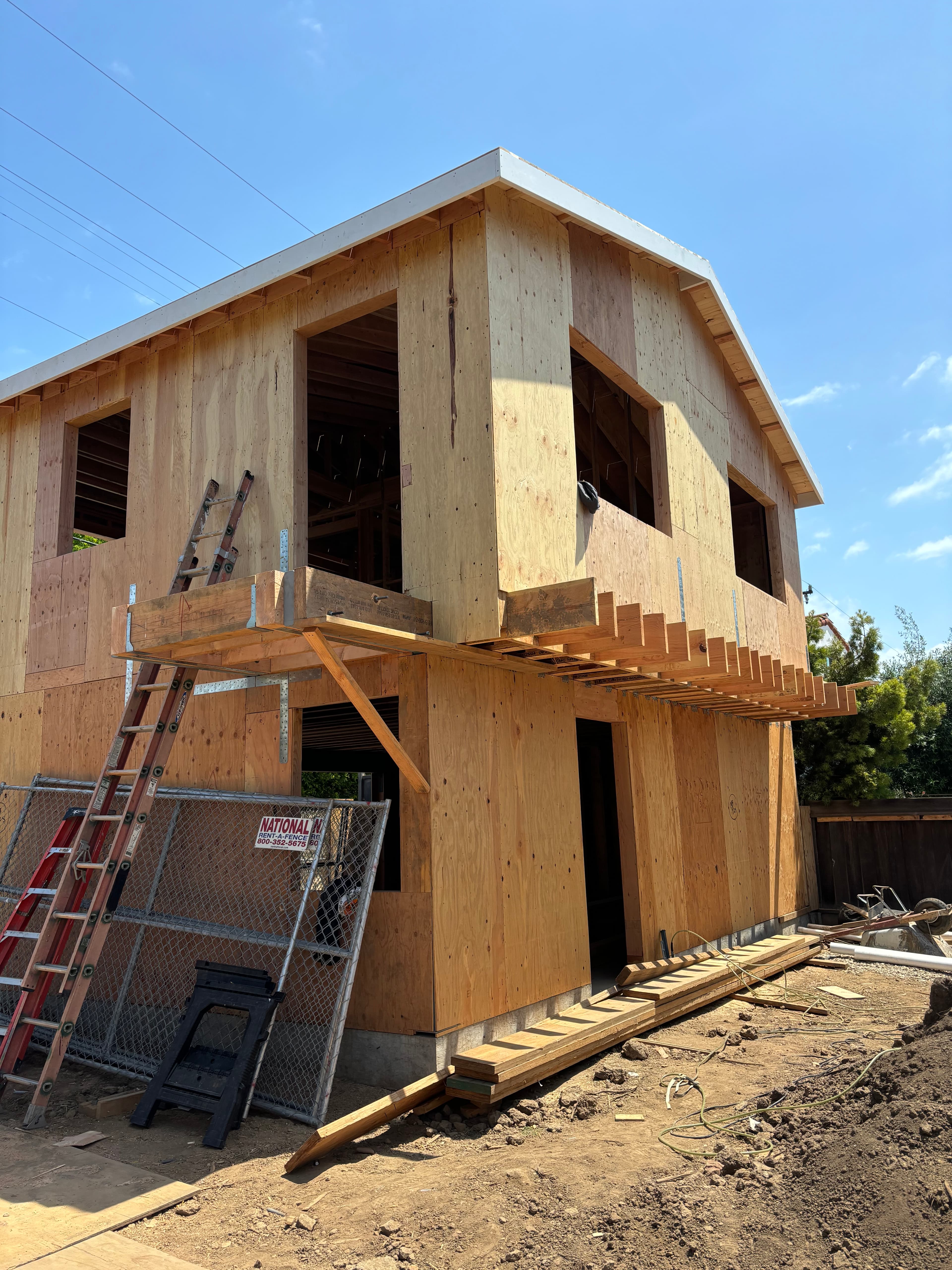 A two-story house under construction with plywood sheathing, a ladder, and a chain-link fence.
