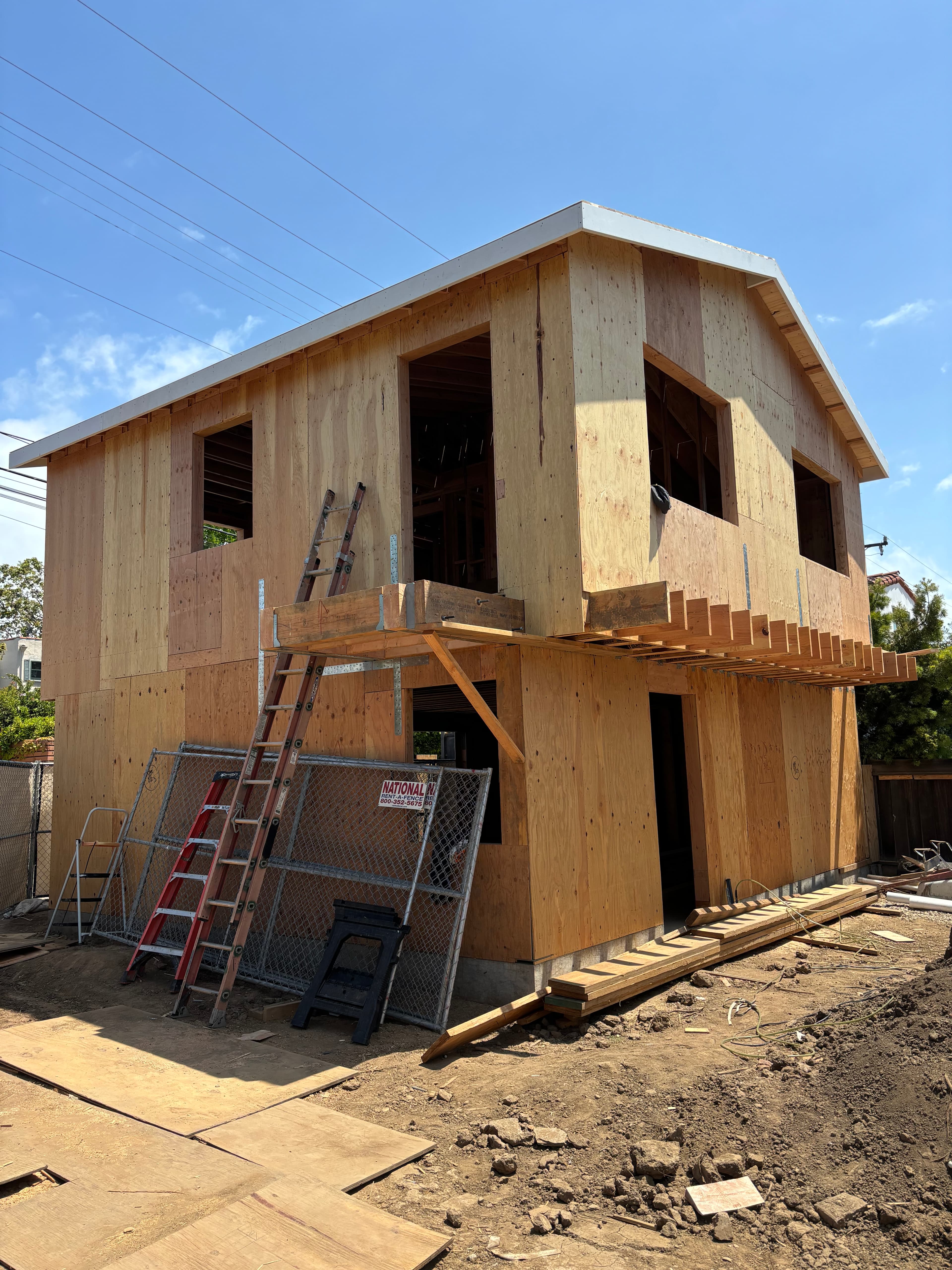 Two-story house under construction with plywood sheathing, ladders, and fencing under a blue sky.