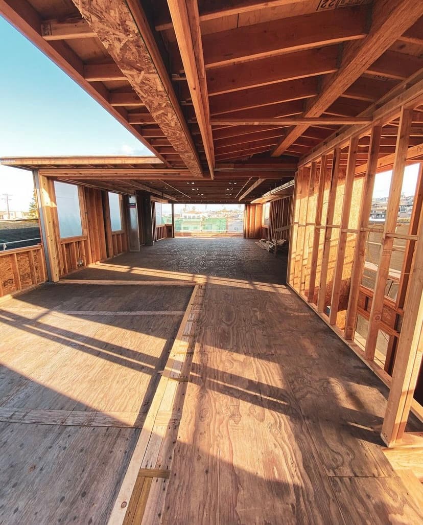 Interior of a wooden building under construction with sunlight casting shadows on plywood floors.