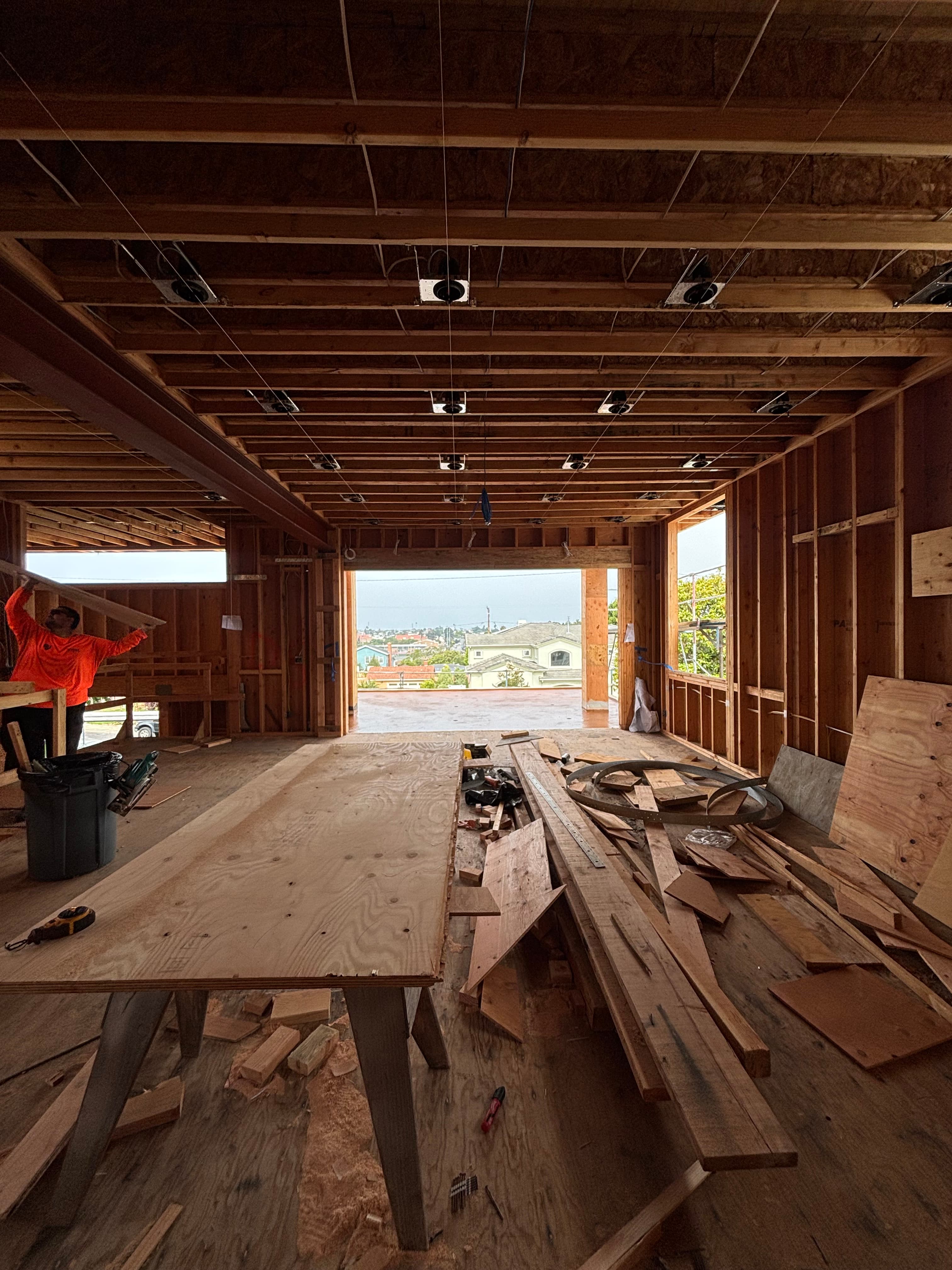 Interior of a house under construction with wooden framing, exposed ceiling joists, and scattered lumber.