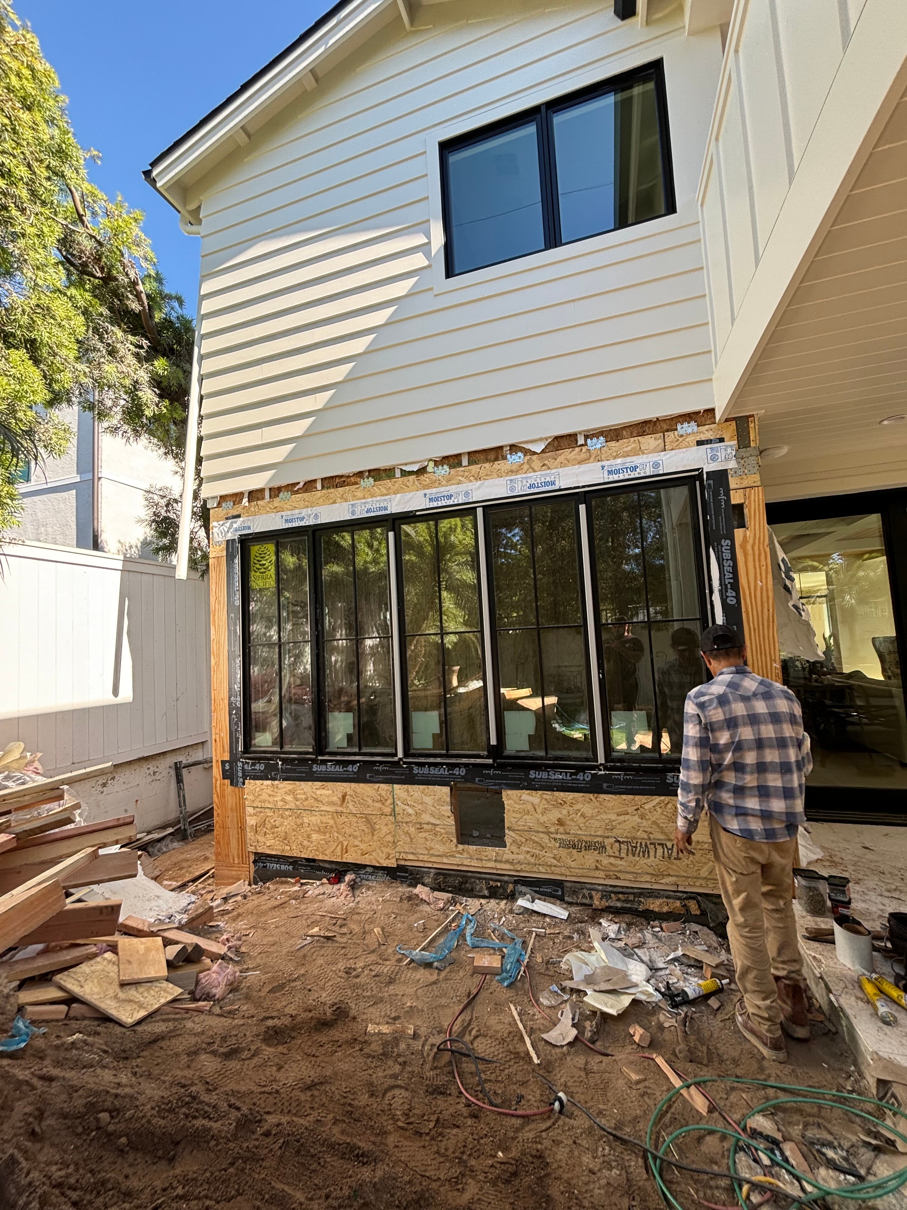 Worker inspecting a large black-framed window installation on a house under construction.